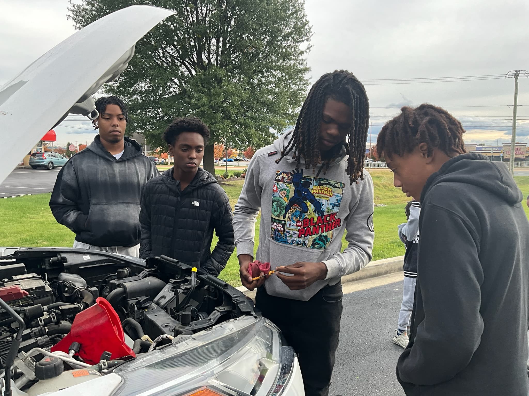 a teen checks the engine oil dipstick while another teen watches and learns